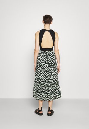 Persona de pie de espaldas, con camiseta negra sin mangas con un gran recorte en la espalda, falda midi estampada y sandalias negras sobre un suelo gris.