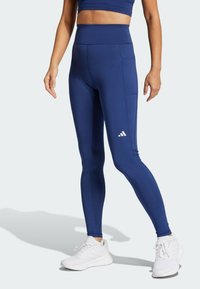Blaue hochtaillierte Leggings mit einer glatten Textur, figurbetontem Design und einem kleinen weißen Logo. Kombiniert mit weißen Sportschuhen.