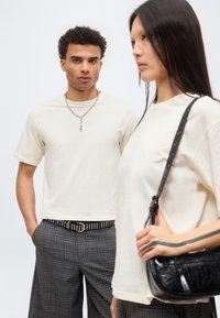 Άνδρας και γυναίκα φοράνε λευκά μπλουζάκια και γκρι καρό παντελόνια· ο άνδρας φοράει αλυσίδα και ζώνη με καρφιά· η γυναίκα κρατάει μια μαύρη τσάντα ώμου.