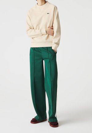 Osoba ubrana w beżową bluzę z logo Lacoste, zielone szerokie spodnie z bocznymi paskami oraz bordowe wsuwane buty, stojąca z jedną ręką w kieszeni.