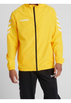 Homme portant une veste zippée jaune avec des bandes chevrons blanches sur les manches et un pantalon noir, tous deux de la marque "hummel."