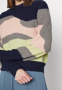 Personne portant un pull multicolore avec des motifs abstraits bleus, gris, roses et vert clair, et un bracelet avec des perles et des perles souriantes.