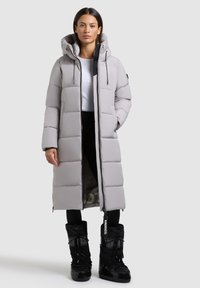 Lång grå puffjacka med huva, med dragkedjestängning och horisontell quiltad design. I kombination med svarta vinterstövlar.