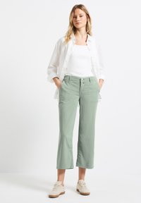 Pantalones cortos de color verde claro con un frente plano, dos bolsillos y un cierre de botón, combinados con una camiseta sin mangas blanca y una camisa de lino blanca.