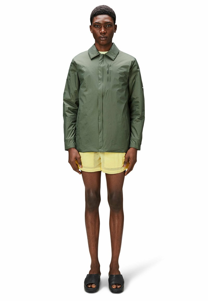 Homme debout portant une veste légère olive, un short jaune et des sandales noires à bout ouvert sur un fond blanc.