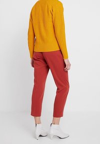 Žlutý žebrovaný svetr, přiléhavý horní díl, kombinovaný s rezavě zbarvenými zúženými kalhotami. Bílé kotníkové boty na zip. Jednoduché bílé pozadí.