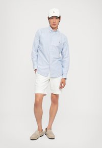 Lichtblauwe gestreepte button-down shirt, witte shorts, grijze instapschoenen en een witte pet met een klein logo, staande tegen een neutrale achtergrond.