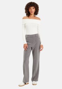 Donna con capelli ricci che indossa un top bianco a spalla scoperta, pantaloni grigi a gamba larga e sneakers bianche, in piedi di fronte a uno sfondo neutro.