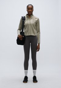 Camisola desportiva de manga comprida verde-claro, leggings cinzentas, ténis pretos e uma pequena mala preta. Tecido liso e elegante com fecho de correr frontal.