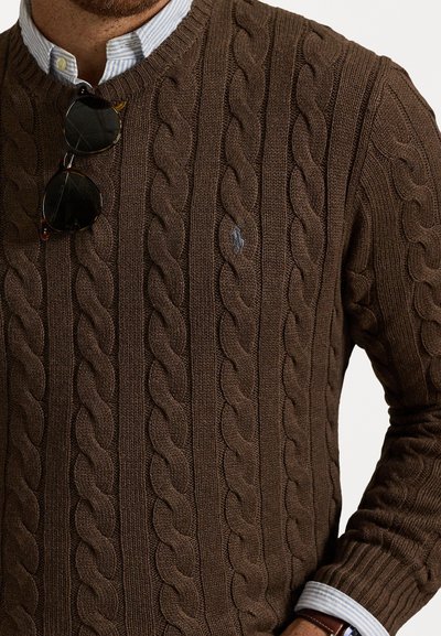 Bruin kabelgebreid sweater met een geribbelde structuur, V-hals en klein geborduurd logo. Draagt over een blauw gestreept overhemd met kraag.