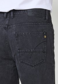 Los jeans de mezclilla gris oscuro presentan un diseño clásico de cinco bolsillos con una pequeña etiqueta de logo en el bolsillo trasero y un acabado de tela texturizada.