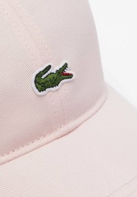 Różowa czapka baseballowa z haftowanym zielonym logo krokodyla. Wykonana z teksturowanej tkaniny, ma zakrzywiony daszek oraz widoczne detale haftu.