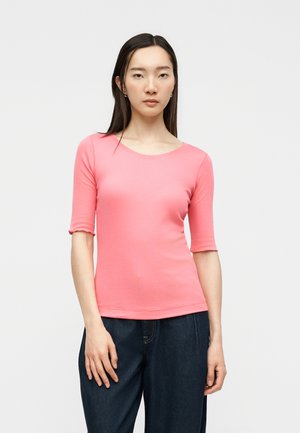 Νεαρή γυναίκα με μακριά σκούρα μαλλιά, που φοράει ροζ τοπ με μανίκια τριών τετάρτων και σκούρα μπλε τζιν, στέκεται μπροστά σε ένα απλό φωτεινό φόντο.