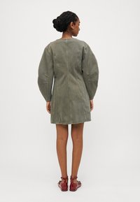 Ελιά πράσινο φόρεμα με εφαρμοστό κορσέ, φούστα που φουσκώνει και υπερμεγέθη μανίκια. Οι ακατάλληλες άκρες και οι λεπτομέρειες ραφής τονίζουν το σχέδιο.