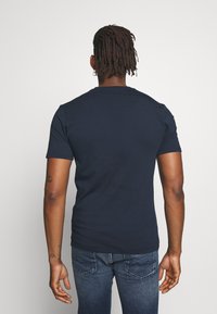 Uomo con dreadlocks che indossa una maglietta blu navy semplice e jeans blu, in piedi di spalle su uno sfondo chiaro e uniforme.