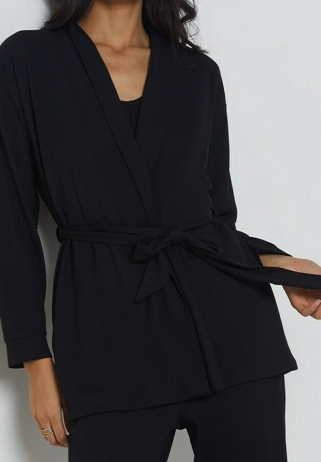 Femme portant une veste noire texturée à nouer à la taille avec des manches longues et un pantalon noir assorti sur un fond uni.