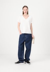 White cotton v-neck t-shirt paired with loose-fit, dark blue denim jeans. Model holds a small beige handbag and wears black sneakers.