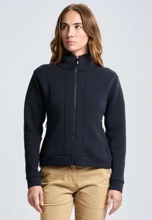 Una mujer luce una chaqueta azul marino oscuro con cremallera y cuello alto, y unos pantalones de color tostado, sobre un fondo liso y claro.