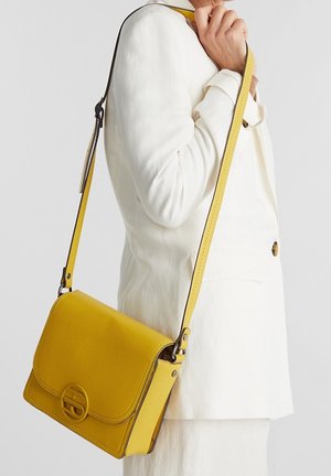 Vrouw draagt een witte blazer en heeft een gele leren schoudertas met een klep en een ronde logo-sluiting.