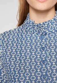 Femme portant un chemisier bleu à motif floral blanc, col montant à volants et boutons recouverts de tissu sur le devant.