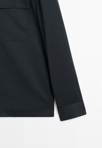 Camicia nera a maniche lunghe con taschino sul petto, texture liscia e polsini dritti. Il tessuto sembra leggero e leggermente elastico.