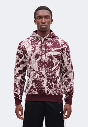Jeune homme portant un sweat à capuche bordeaux et blanc à motif abstrait avec poche avant et short noir Nike, debout devant un fond uni.