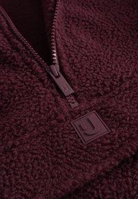 Burgundi színű fleece pulóver negyed cipzárral, texturált anyaggal, tónusos cipzárral és egy kis domború logóval.