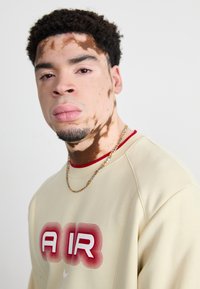 Μπεζ φούτερ με έντονο κόκκινο και λευκό γραφικό "AIR", κόκκινη λαιμόκοψη και χρυσή αλυσίδα ως αξεσουάρ. Υφή επιδερμίδας με διακριτά μοτίβα.