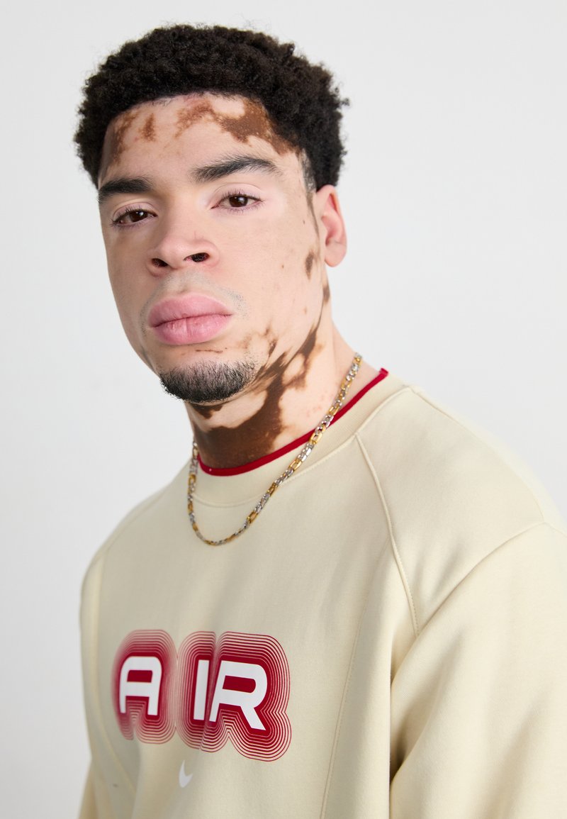Beige Sweatshirt mit auffälligem roten und weißen "AIR"-Grafik, rotem Kragen und goldener Halskette; strukturierten Haut mit ausgeprägten Mustern.