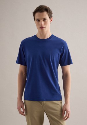 Giovane uomo in piedi contro uno sfondo semplice, indossa una maglietta blu a maniche corte e pantaloni beige, guarda avanti con un'espressione neutra.