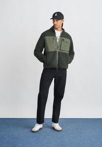 Πράσινο φλις και ύφασμα ζακέτα με κλείσιμο φερμουάρ και τσέπη στο στήθος, συνδυασμένο με σκούρα παντελόνια και λευκά αθλητικά παπούτσια. Το μοντέλο φοράει ένα μαύρο καπέλο.