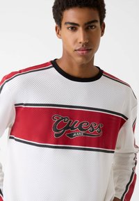 Biała koszula z siateczki z długim rękawem, z czerwono-czarnymi paskami, z wyraźnym logo "Guess Jeans" na piersi w kolorze czarno-białym.