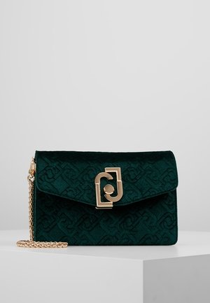 Pochette en velours vert foncé avec motif embossé, fermoir géométrique doré et chaîne dorée sur une surface blanche.