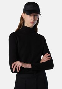 Schwarzer gerippter Rollkragenpullover mit langen Ärmeln, kombiniert mit einer schwarzen Mütze mit einem kreisförmigen Logo. Der Stoff wirkt weich und glatt.