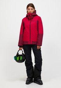 Rød og kastanjebrun vandtæt skijakke med lynlåslukning, justerbar hætte og sorte skibukser. Inkluderer en sort hjelm med grøn visir.