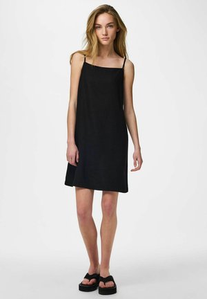 Junge Frau mit langen blonden Haaren trägt ein ärmelloses schwarzes Kleid und schwarze Plateausandalen, steht vor einem schlichten Hintergrund.