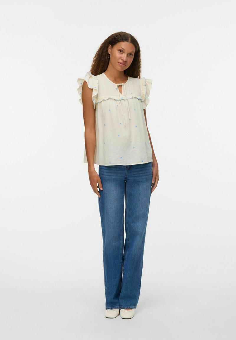 Top bianco con maniche arricciate e un fiocco al collo, abbinato a jeans azzurri chiari. Il top presenta piccole macchie blu.