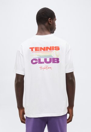 Άνδρας με την πλάτη προς τα έξω φοράει λευκό μπλουζάκι με το κείμενο "TENNIS CLUB", πολύχρωμες γραμμές, μια τενιστική μπάλα και μωβ σορτς.