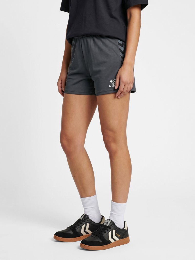 Personne portant un short de sport noir, des baskets noires avec des chevrons blancs et des chaussettes blanches, debout sur un fond blanc uni.