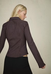 Cămașă ajustată de un burgund închis, cu dungi verticale subțiri, mâneci lungi, guler tăiat și un tiv curbat, confecționată dintr-un material neted.