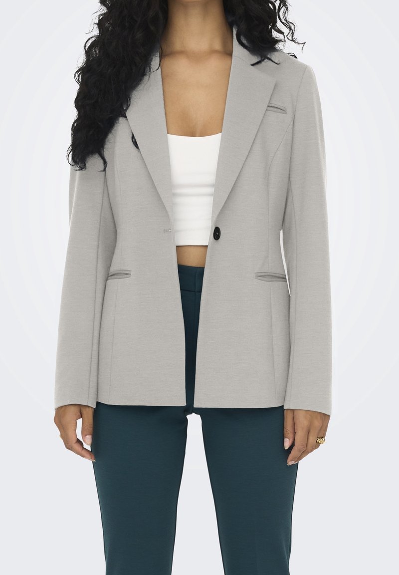 Femme portant un blazer gris clair, un top court blanc et un pantalon bleu sarcelle foncé, vue du cou à mi-cuisse, les mains détendues le long du corps.