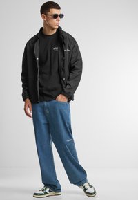Svart zip-up jakke, svart t-skjorte, slitte blå jeans og grønne og hvite joggesko. Tilbehør inkluderer mørke solbriller.