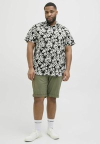 Schwarzes Kurzarmhemd mit weißen Blattdruck, kombiniert mit olivgrünen Shorts, weißen Sneakers und Socken mit Logo. Das Modell steht vor einem neutralen Hintergrund.