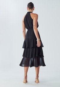 Nainen, joka pukeutunut mustaan, kerrokselliseen hihattomaan mekkoon, jossa on korkea kaulus, ja nudenvärisiin korkokenkiin, seisoo selin yksiväristä taustaa vasten.