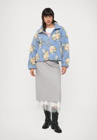 Sinine fliisjakke lillemustriga, millel on tõmbluku sulgemine, ümar krae ja elastsed mansetid. Kandmise all on halli pliiatsseelik.