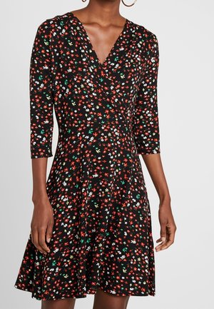 Femme portant une robe noire à petites fleurs rouges, blanches et vertes, manches trois-quarts et encolure en V.