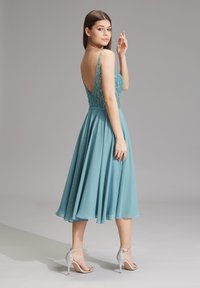 Aqua-blaues Kleid mit besticktem Oberteil, tiefem Rückenausschnitt und ausgestelltem Chiffonrock. Das Model trägt silberne High Heels.