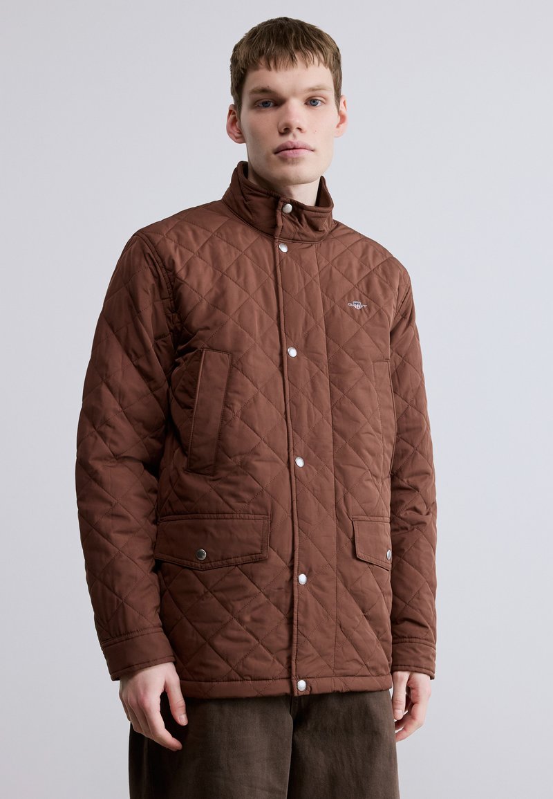 Veste matelassée marron avec boutons pression, col montant et deux poches avant, présentant une texture lisse et une coupe ajustée.