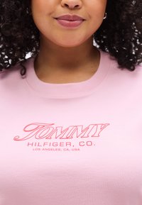 Ανοιχτό ροζ βαμβακερό t-shirt με στρογγυλή λαιμόκοψη, που διαθέτει έντονη κόκκινη κεντητή επιγραφή "TOMMY HILFIGER, CO." με λεπτ ομέρειες τοποθεσίας από κάτω.