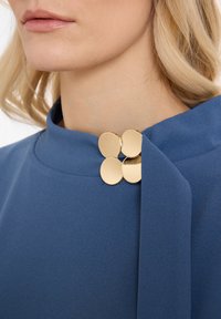 Tissu bleu avec une texture lisse, présentant un col haut et trois accents ronds dorés en tant que fermeture décorative, positionnés au niveau du cou.